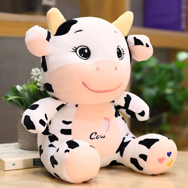 peluche-vache-jouet-enfant-bebe-cadeau-noel-3a Une peluche de vache blanche et noire avec des yeux noirs et un sourire brodé. La peluche est assise sur une table en bois.