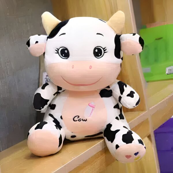 peluche-vache-jouet-enfant-bebe-cadeau-noel-4a Peluche vache noire et blanche avec un biberon sur son ventre, assis sur une étagère en bois.