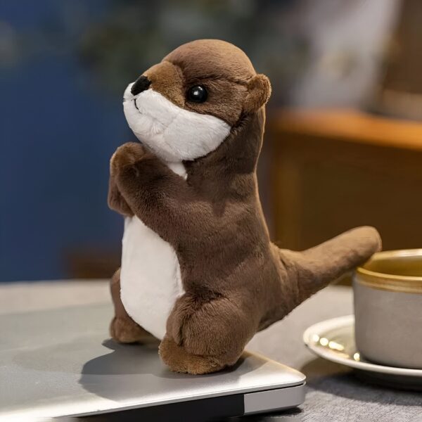 Une peluche de loutre debout sur un ordinateur portable avec une tasse de café sur une soucoupe à côté.