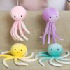 Quatre peluches poulpes de différentes couleurs : rose, violet, jaune et bleu. Les poulpes ont tous des sourires amicaux et des tentacules blancs et moelleux.