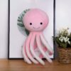 Une peluche de poulpe rose avec des yeux noirs et un sourire, posée sur une table en bois.