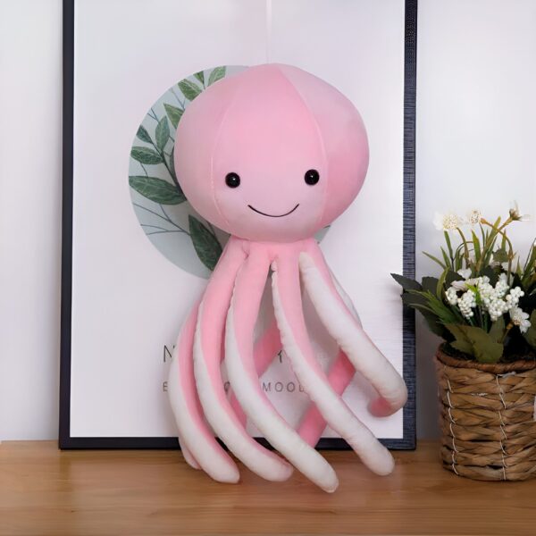 Une peluche de poulpe rose avec des yeux noirs et un sourire, posée sur une table en bois.